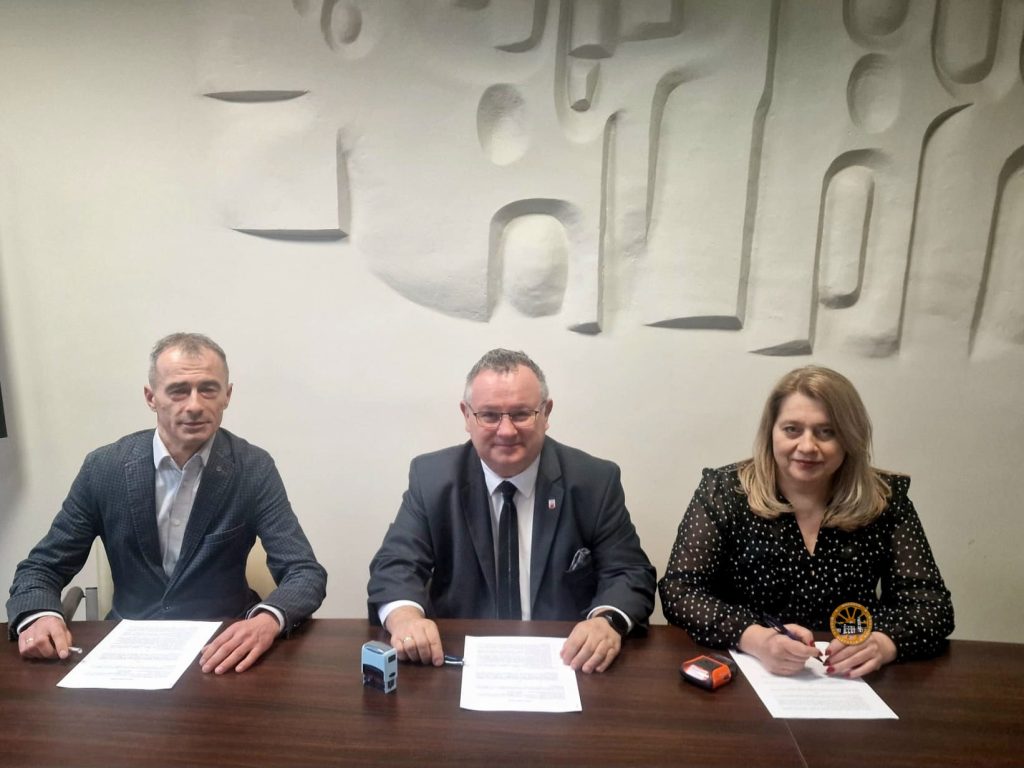 Trzy osoby siedzą przy stole podczas oficjalnego podpisywania dokumentów. W centrum znajduje się mężczyzna w garniturze i krawacie, po jego lewej stronie siedzi mężczyzna w marynarce, a po prawej kobieta w czarnej bluzce w kropki. Każda z osób ma przed sobą dokument i długopis, na stole widoczne są także pieczątki, a w tle znajduje się jasna ściana z dekoracyjnym wzorem.