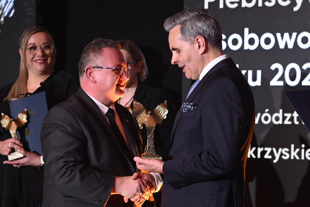 Dwóch mężczyzn w garniturach stoi na scenie podczas uroczystości i ściska sobie dłonie, jeden z nich wręcza drugiemu statuetkę. W tle widoczna jest kobieta trzymająca nagrodę oraz ekran z napisem dotyczącym plebiscytu.