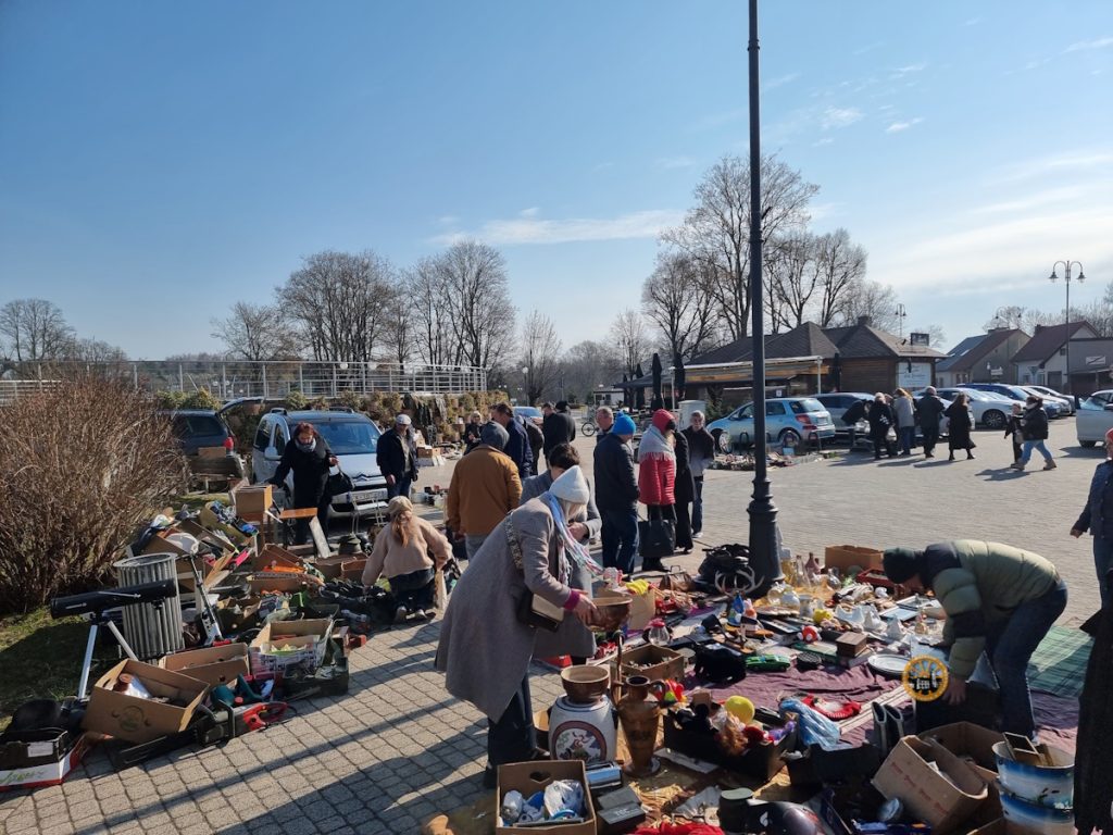 Wypełnione słońcem, szerokie ujęcie tętniącego życiem pchlego targu na świeżym powietrzu, zlokalizowanego na wybrukowanym placu. Na pierwszym planie na ziemi i w kartonowych pudełkach rozłożone są liczne starocia, w tym ceramika, narzędzia, wazy, figurki, naczynia kuchenne, a po lewej stronie teleskop na statywie. Kilka osób przegląda towary. Centralnie kobieta w szarym płaszczu i beżowej czapce pochyla się nad ceramiczną miską, a po prawej mężczyzna w oliwkowej kurtce kuca, badając przedmioty. W tle widać parking z zaparkowanymi samochodami, w tym srebrnego vana, pawilon restauracyjny z tarasem i gołymi drzewami pod jasnym, błękitnym niebem z lekkimi chmurami. Atmosfera jest busy i pełna odkryć.