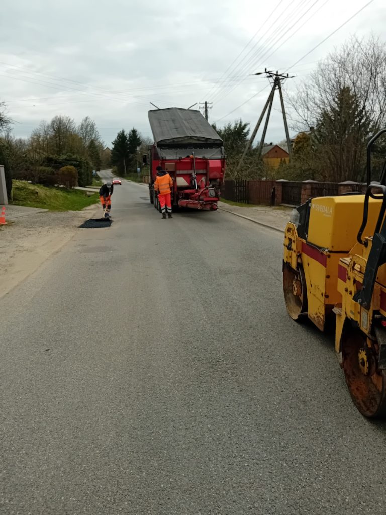 Widok z poziomu drogi z tyłu na aktywne prace drogowe na wiejskiej drodze. Po prawej stronie widać fragment żółtego walca drogowego marki Volvo. Na środku drogi stoi duża czerwona rozkładarka asfaltu, a przy niej pracuje dwóch robotników w pomarańczowych kurtkach i kaskach. Jeden używa łopaty do wygładzania świeżej, czarnej plamy nowego asfaltu. Wzdłuż drogi ciągną się ogrodzenia i drzewa. W oddali widać nadjeżdżający mały czerwony samochód.