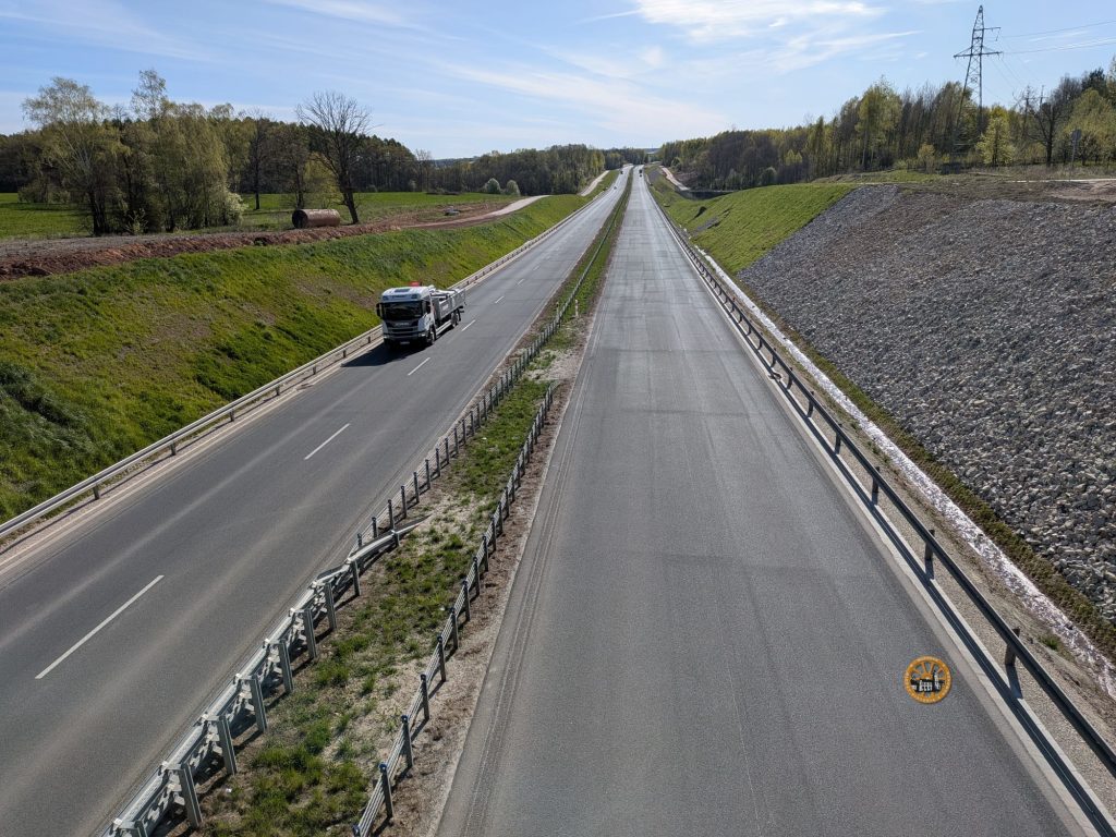 Widok z góry na dwujezdniową drogę ekspresową ciągnącą się w głąb krajobrazu. Na lewej jezdni jedzie biała ciężarówka wywrotka. Jezdnie oddzielone trawą, po prawej kamienny nasyp. W oddali lasy i linia energetyczna pod bezchmurnym niebem.