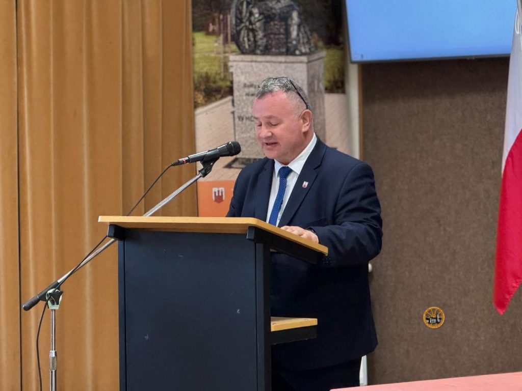 Oto opis alt dla zdjęcia:

Zdjęcie średniego planu mężczyzny w ciemnogranatowym garniturze, białej koszuli i wzorzystym niebieskim krawacie, przemawiającego zza ciemnego pulpitu z drewnianym blatem. Ma krótkie, szpakowate włosy i patrzy lekko w dół, mówiąc do dynamicznego mikrofonu zamontowanego na statywie. Za nim znajdują się złote zasłony, baner przedstawiający zabytkowy pomnik i herb Gminy Wąchock, oraz flaga Polski. W prawym dolnym rogu widnieje małe, okrągłe logo.