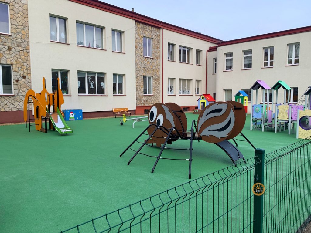Nowoczesny plac zabaw z zieloną, bezpieczną nawierzchnią, zlokalizowany na dziedzińcu piętrowego budynku szkoły lub przedszkola. Na pierwszym planie widoczna jest duża konstrukcja wspinaczkowa w kształcie pająka.