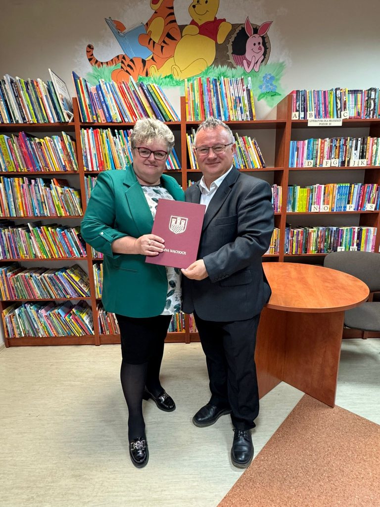 Kobieta w zielonej marynarce i mężczyzna w ciemnym garniturze pozują w bibliotece, trzymając wspólnie bordową teczkę z herbem Gminy Wąchock. W tle widać regały z książkami oraz ścienne malowidło przedstawiające postacie z bajki o Kubusiu Puchatku.