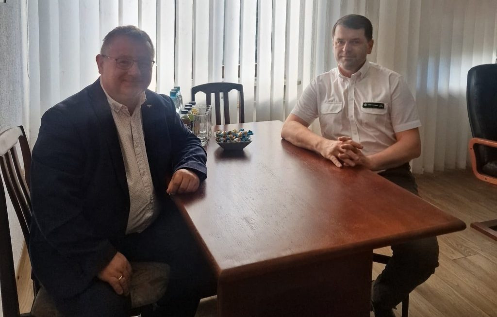 Fotografia przedstawia dwóch mężczyzn siedzących przy dużym, drewnianym stole w jasnym pomieszczeniu biurowym. Mężczyzna po lewej stronie jest uśmiechnięty, nosi okulary, jasną wzorzystą koszulę i ciemnogranatową marynarkę. Mężczyzna po prawej stronie ma na sobie białą koszulę z krótkim rękawem (z widocznym ciemnym identyfikatorem na piersi) i ciemne spodnie, a jego dłonie są splecione na blacie. Na stole między nimi stoi miseczka z cukierkami oraz małe butelki z napojami i szklanki. W tle znajduje się okno zasłonięte jasnymi, pionowymi żaluzjami.