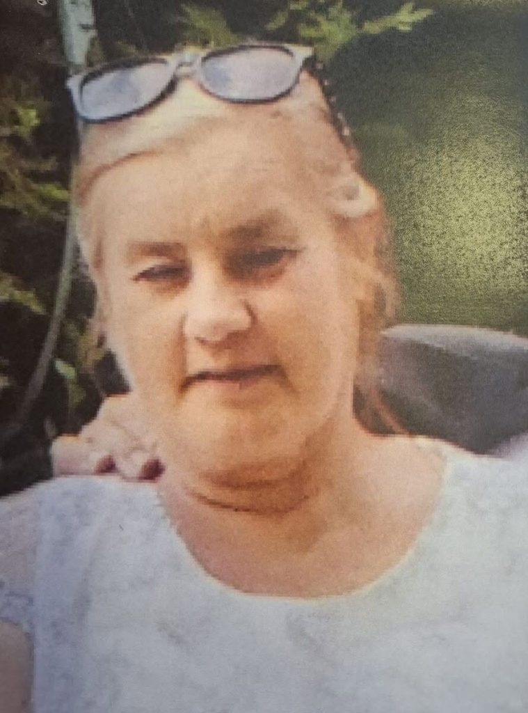Zdjęcie portretowe starszej kobiety z delikatnym, neutralnym wyrazem twarzy. Kobieta ma jasne, zaczesane do tyłu włosy, na których opiera ciemne okulary przeciwsłoneczne. Ubrana jest w jasną bluzkę z drobnym, jasnym wzorem. Tło za nią jest rozmyte, utrzymane w ciemnozielonej, naturalnej kolorystyce, przypominającej liście drzew. Fotografia jest lekko nieostra i ma widoczną strukturę rastra lub odbicia, co sugeruje, że jest to zdjęcie wydrukowanej wcześniej odbitki lub ekranu.