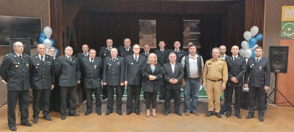Ustawione do zdjęcia grupowego kilkanaście osób, w większości mężczyzn w ciemnych, galowych mundurach strażackich z odznakami, stojących na scenie. W środku pierwszego rzędu stoi kobieta w czarnym garniturze, obok niej mężczyźni w cywilnych ubraniach, w tym jeden w piaskowym mundurze specjalnym. Tło stanowią ciemne kurtyny, dwa plakaty (jeden z napisem "WĄCHOCK") oraz białe i błękitne balony po bokach.