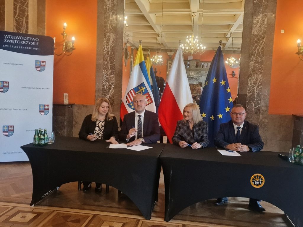 Zdjęcie przedstawia cztery osoby siedzące obok siebie za połączonymi stołami, przykrytymi czarnym materiałem, w eleganckiej sali o pomarańczowych ścianach i zdobionych, marmurowych filarach. Od lewej strony siedzą: kobieta w czarno-białej wzorzystej bluzce oraz łysy mężczyzna w ciemnym garniturze – oboje trzymają długopisy i są w trakcie podpisywania dokumentów leżących na blacie. Obok nich siedzą uśmiechnięta kobieta w ciemnej marynarce w kratę oraz mężczyzna w okularach, ubrany w granatowy garnitur, którzy opierają dłonie na stole.
Tuż za uczestnikami spotkania stoją trzy flagi (od lewej): województwa świętokrzyskiego, Polski oraz Unii Europejskiej. Z lewej strony kadru widoczny jest pionowy biały baner z herbem i napisami „WOJEWÓDZTWO ŚWIĘTOKRZYSKIE”. Na stole przed osobami stoją szklanki oraz małe butelki z wodą mineralną. W tle widoczne jest duże lustro, w którym odbija się bogato zdobiony sufit z kryształowymi żyrandolami. W prawym dolnym rogu zdjęcia, na czarnym tle obrusu, znajduje się niewielkie, okrągłe, pomarańczowe logo.
