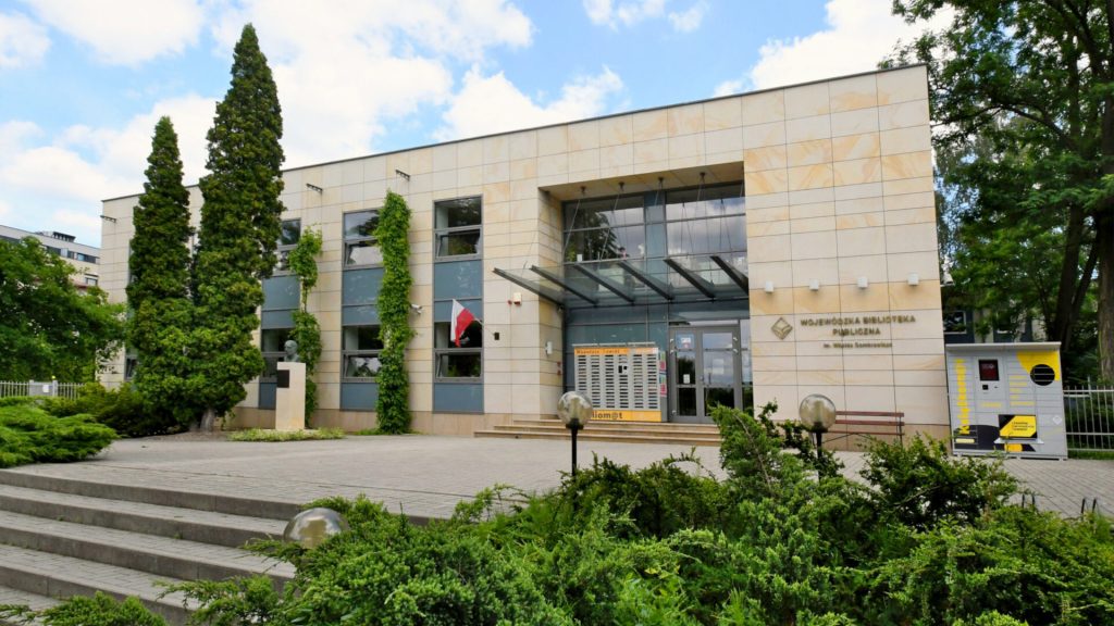 Fotografia przedstawia dwupiętrowy budynek o nowoczesnej bryle, obłożony jasnymi, piaskowymi płytami. Jest to siedziba Wojewódzkiej Biblioteki Publicznej im. Witolda Gombrowicza, o czym informuje elegancki napis na elewacji po prawej stronie. Centralną część budynku stanowi szerokie, przeszklone wejście osłonięte płaskim, szklanym zadaszeniem. Po lewej stronie od drzwi wisi polska flaga, a przestrzenie pomiędzy sąsiadującymi oknami obrastają gęste, zielone pnącza.
Przed budynkiem znajduje się rozległy, wybrukowany plac, do którego prowadzą szerokie, łagodne schody. Na placu, po lewej stronie, zlokalizowano pomnik w formie popiersia na jasnym cokole, obok którego rosną wysokie, smukłe drzewa iglaste. Z prawej strony, nieopodal wejścia, ustawiono żółto-szare urządzenie przypominające paczkomat z napisem „Książkomat”. Na pierwszym planie widoczne są obfite, zielone krzewy ozdobne. Nad budynkiem rozpościera się błękitne niebo z białymi obłokami, a cała scena jest jasno oświetlona światłem słonecznym.
