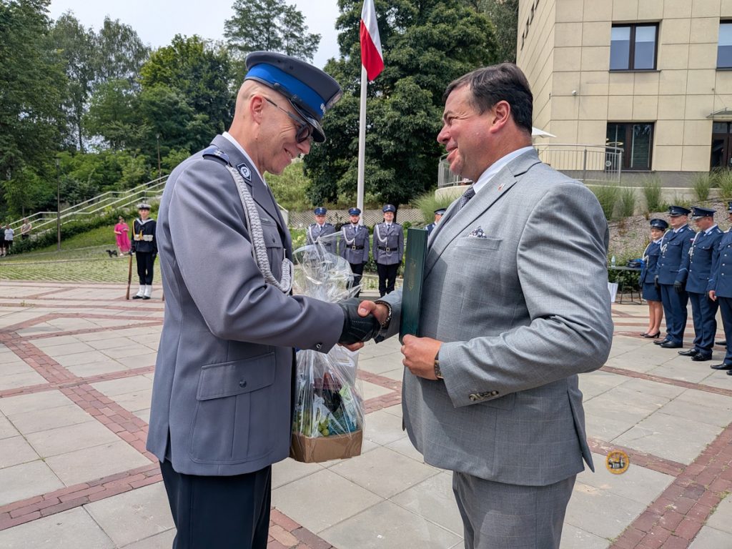 Uroczyste wręczenie upominku z okazji Święta Policji w Starachowicach. Na pierwszym planie, na placu przed budynkiem i w otoczeniu zieleni, zastępca Burmistrza Norbert Dudek (w szarym garniturze, trzymający zielony folder) ściska dłoń aspirantowi Łukaszowi Jasikowi (w pełnym mundurze galowym policji, z czapką i w okularach przeciwsłonecznych). Funkcjonariusz w drugiej ręce trzyma kosz prezentowy owinięty folią. W tle widoczna jest asysta honorowa policjantów w mundurach, maszt z flagą Polski oraz budynek urzędu.