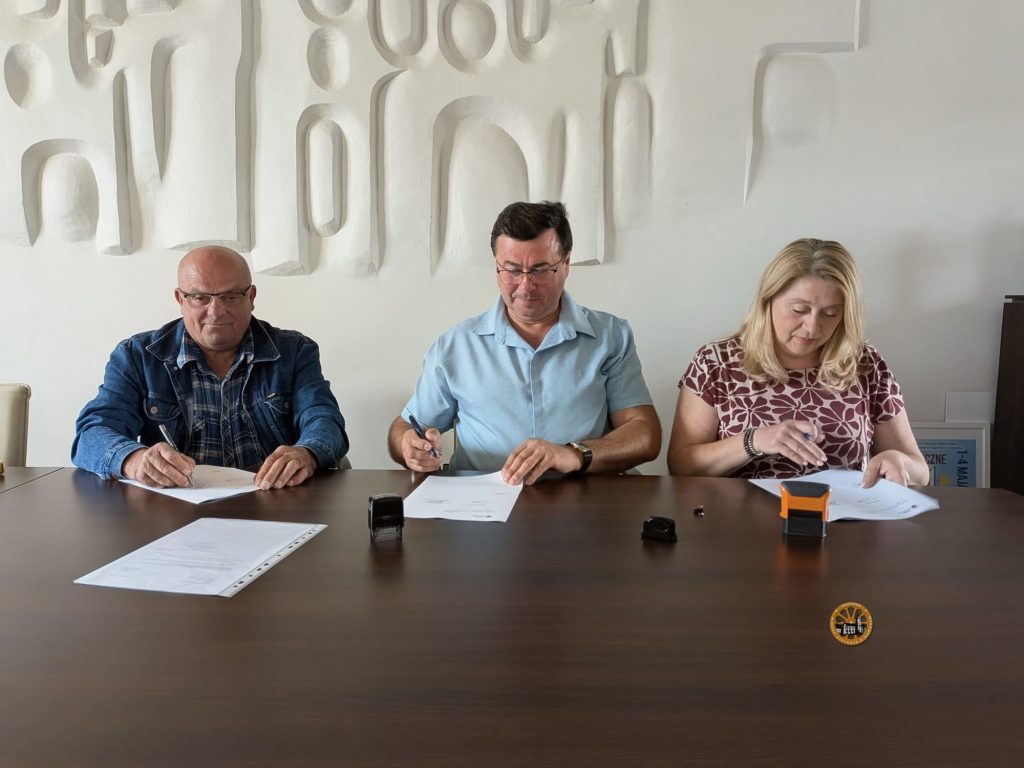 Uroczysta fotografia przedstawiająca ceremonię podpisania umowy. Trzy osoby siedzą obok siebie przy dużym, ciemnym, drewnianym stole konferencyjnym. Po lewej stronie, starszy, łysy mężczyzna w ciemnoniebieskiej kurtce dżinsowej i koszuli w kratę podpisuje dokument długopisem. W środku, mężczyzna w średnim wieku, w okularach i jasnoniebieskiej koszuli z krótkim rękawem, również składa podpis pod papierem, spoglądając w dół. Po prawej stronie, kobieta z blond włosami, ubrana w wzorzystą, czerwoną bluzkę z krótkim rękawem, podpisuje swój egzemplarz dokumentu. Każda z osób trzyma długopis. Na stole widoczne są stosy dokumentów, długopisy oraz dwa różne rodzaje małych podkładek do stempli. Tło stanowi czysta, biała ściana z unikalną, nowoczesną rzeźbą reliefową o zakrzywionych, łukowatych kształtach.