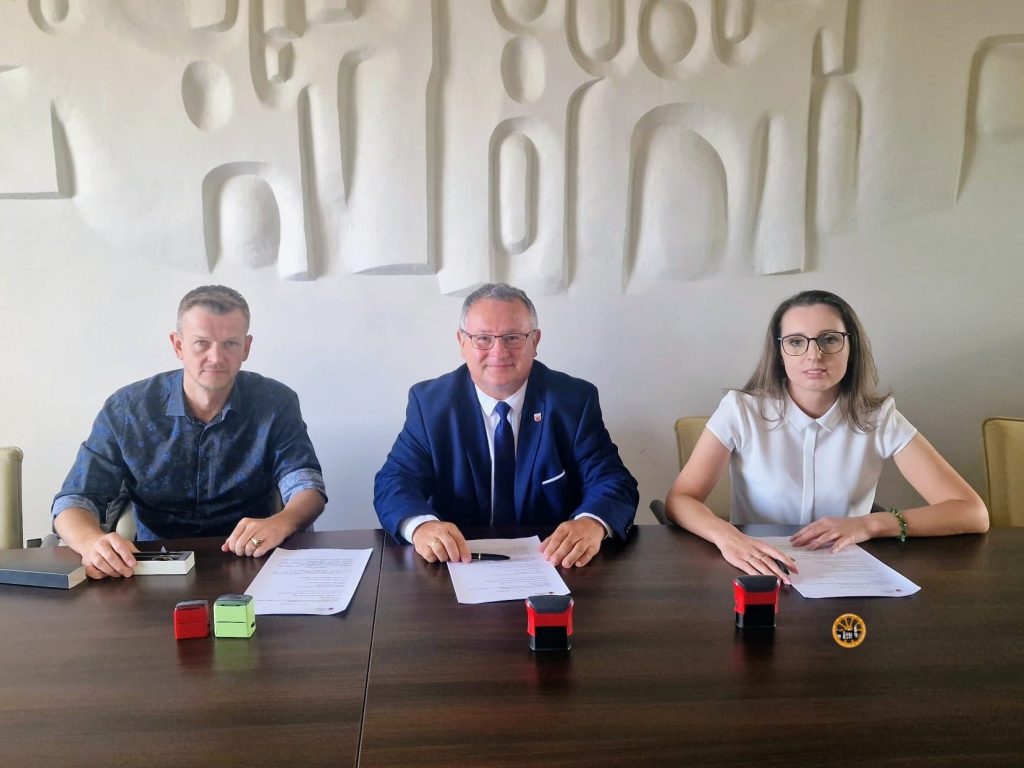 Fotografia oficjalnego spotkania trzech osób przy stole konferencyjnym, dokumentująca moment po lub w trakcie podpisywania umowy. Centralnie siedzi mężczyzna w garniturze i okularach, po jego prawej kobieta w białej bluzce i okularach, a po lewej mężczyzna w niebieskiej koszuli we wzory. Wszyscy patrzą w stronę aparatu. Przed nimi na stole leżą dokumenty, długopisy i małe urządzenia biurowe (w tym dwa urządzenia do podpisu cyfrowego). W tle biała ściana z abstrakcyjnym reliefem.