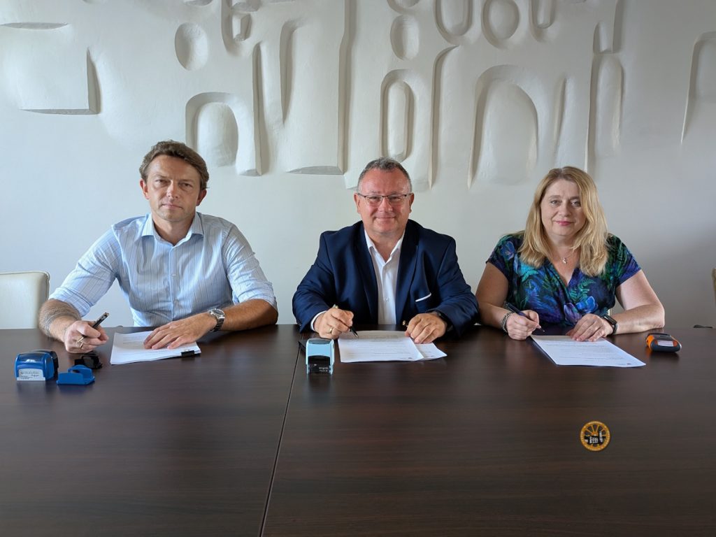  opis alt

Fotografia przedstawia trzy osoby siedzące obok siebie za długim, ciemnym stołem na tle jasnej ściany ozdobionej przestrzenną, geometryczną płaskorzeźbą. Od lewej strony siedzą: mężczyzna w błękitnej koszuli w prążki z podwiniętymi rękawami, Burmistrz Miasta i Gminy Wąchock Robert Janus ubrany w ciemną marynarkę i białą koszulę oraz jasnowłosa kobieta w niebiesko-zielonej bluzce w roślinne wzory. Wszystkie trzy osoby trzymają w dłoniach długopisy opuszczone na leżące przed nimi dokumenty, uśmiechając się do obiektywu w momencie podpisywania umów. Na blacie stołu, przed każdą z osób, znajdują się różnokolorowe pieczątki biurowe.