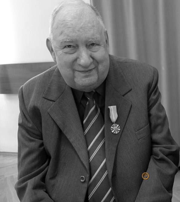 Czarno-białe zdjęcie starszego, siwiejącego mężczyzny o pogodnej twarzy, patrzącego prosto w obiektyw z ciepłym uśmiechem. Ubrany jest w marynarkę, ciemną koszulę i krawat w ukośne paski. Na lewej klapie jego marynarki (z perspektywy widza) przypięty jest medal na paskowanej wstążce. Tło stanowi proste wnętrze z fragmentem drewnianego mebla po lewej i pionową zasłoną po prawej. W prawym dolnym rogu widoczny jest mały, okrągły, pomarańczowy znak wodny.