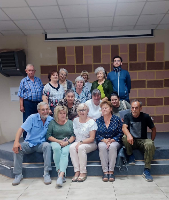 Grupa kilkunastu osób, kobiet i mężczyzn w różnym wieku, pozująca do grupowego zdjęcia w świetlicy. Część z nich siedzi na brzegu niewysokiej sceny, pozostali stoją z tyłu, uśmiechając się do obiektywu.