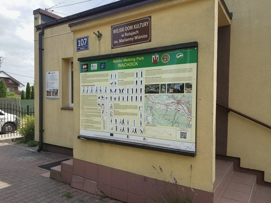 Zdjęcie przedstawia wejście do budynku Wiejskiego Domu Kultury w Ratajach. Na ścianie wisi duża tablica informacyjna z napisem "Nordic Walking Park WĄCHOCK", zawierająca mapę i instrukcje ćwiczeń. Powyżej znajduje się mniejszy szyld: "WIEJSKI DOM KULTURY w Ratajach im. Marianny Wiśnios". Po lewej stronie widoczny jest numer domu 107. Schody prowadzą do wejścia.