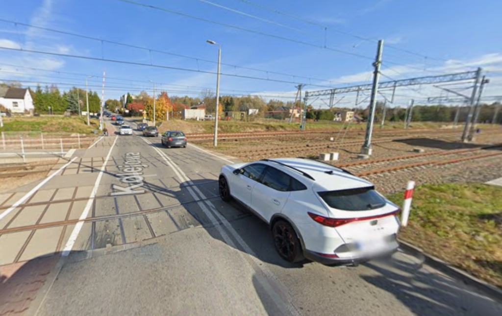 Obraz przedstawia widok z poziomu ulicy na wielotorowy przejazd kolejowy (level crossing) pod bezchmurnym niebem, patrząc wzdłuż torów. Po prawej stronie na poboczu zaparkowany jest biały crossover SUV, częściowo zasłaniając widok. Na lewo od niego widać jezdnię, gdzie inne samochody czekają przed przejazdem. Przejazd posiada wiele równoległych torów, napowietrzne linie trakcyjne i gantry dla pociągów elektrycznych. Na far left, widoczne jest przejście dla pieszych ze specjalnymi żółto-czerwonymi płytkami i napisem "Kolejowa" na asfalcie. Sygnalizacja przejazdu z otwartymi szlabanami znajduje się po prawej stronie. W tle widać drzewa z jesiennymi barwami i kilka budynków.