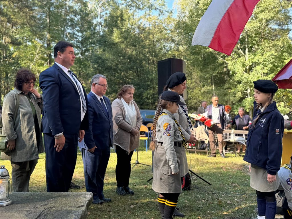 Przedstawiciele władz samorządowych oraz harcerki w mundurach stojący na baczność podczas plenerowych uroczystości patriotycznych. Z boku powiewa polska flaga, a na pierwszym planie widoczna jest kamienna płyta z postawionym zniczem