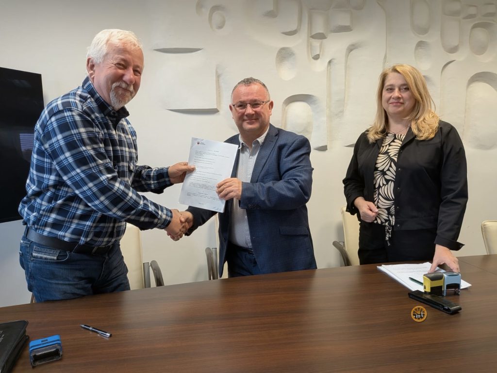 Trzy osoby w jasnym pomieszczeniu podczas uścisku dłoni po podpisaniu dokumentu przy drewnianym stole. Mężczyzna w koszuli w kratę oraz mężczyzna w marynarce trzymają razem umowę i podają sobie ręce, a obok nich stoi uśmiechnięta kobieta w czarnej marynarce. Na stole leżą pieczątki i dokumenty, a w tle znajduje się dekoracyjna, jasna ściana