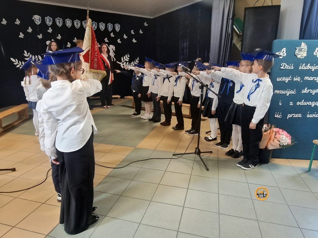 Fotografia przedstawia moment ślubowania pierwszoklasistów w szkolnej sali gimnastycznej. Wzdłuż prawej ściany stoi rząd dzieci w strojach galowych – białych koszulach, ciemnych spodniach i spódniczkach oraz w niebieskich biretach na głowach. Większość z nich ma wyciągnięte do przodu prawe ręce w geście ślubowania, patrząc w lewą stronę, gdzie stoi poczet sztandarowy ze sztandarem szkoły oraz starsze uczennice. Na pierwszym planie, tyłem i bokiem do obiektywu, widać dwoje kolejnych pierwszoklasistów, również w galowych strojach i biretach, z uniesionymi rękami. Po prawej stronie kadru znajduje się niebieska tablica z białymi napisami: „Ślubowanie”, „Dziś ślubuję uczyć się, marzyć, rozwijać skrzydła i z odwagą iść przez świat”. Przed nią, na podłodze, stoi bukiet kwiatów w papierowej owijce. W tle widać ciemną ścianę udekorowaną papierowymi motylami oraz napisem „ŚLUBOWANIE”, a także fragmenty kurtyny i drewniane ławki gimnastyczne. Na pierwszym planie stoi mikrofon na statywie z poprowadzonym po kafelkach kablem. W prawym dolnym rogu zdjęcia umieszczono małe, okrągłe logo z herbem Wąchocka.
